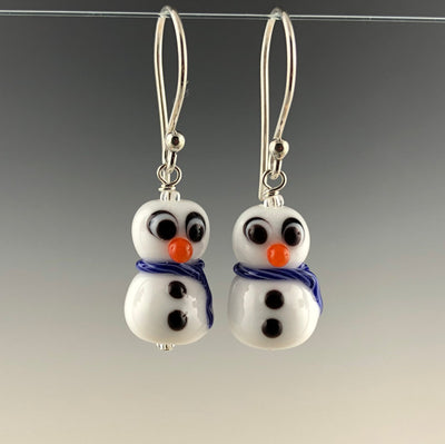 Petite snowmen earrings made of white glass with black and white eyes that look surprized. Each has 2 black buttons down their fronts and orange noses. They have blue and white glass scarves wrapped around their necks. They are on sterling silver ear wires. Zoom ready!
