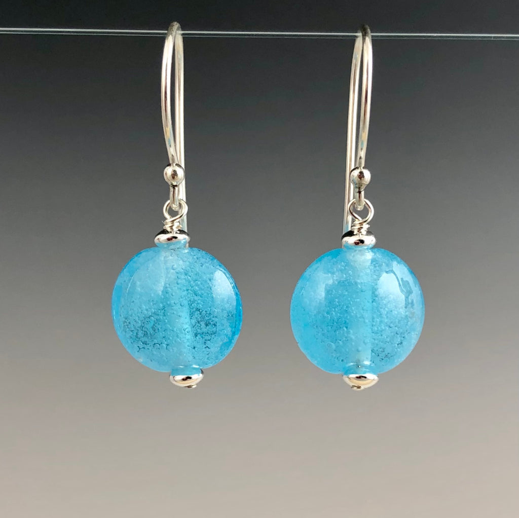 Aquamarine handmade glass beads with tiny air bubbles. Each bead is lentil in shape and dangles from sterling silver Bali ear wires with small silver saucer beads on either side of the glass bead.