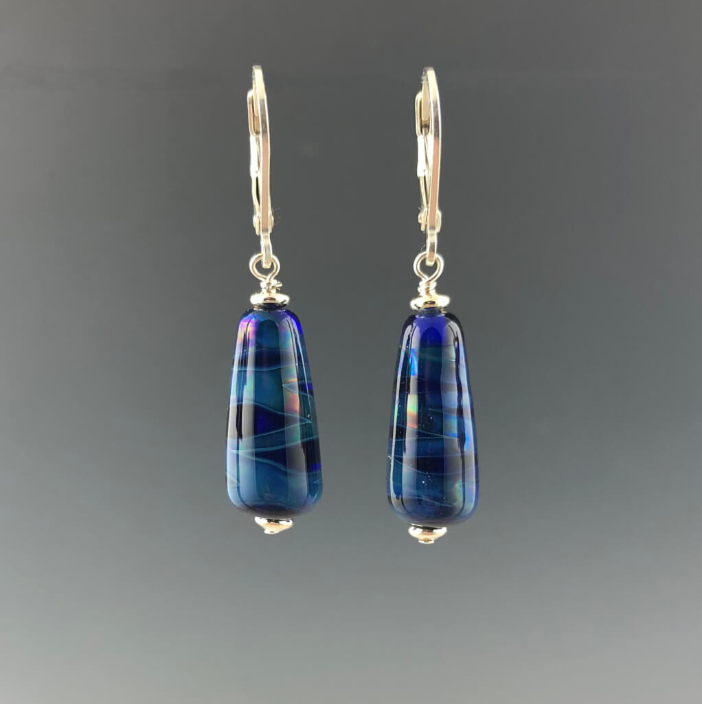Tapered cobalt blue with shimmering light blue lines wrapped around beads dangle from silver leverbacks with small silver saucer beads on top and bottom of the earrings.