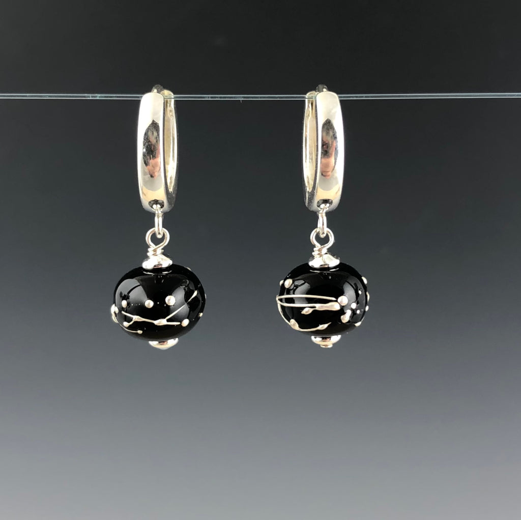 Black Constellation Hoop Earrings by Becky Congdon are handmade black glass beads with fine silver lines and dots wrapped around the beads. Each earring has a modern hinged hoop which has the bead dangling with silver saucer beads on the top and bottom of the glass bead.