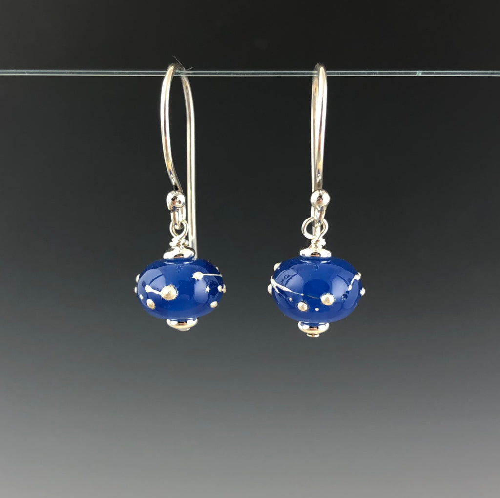 Luna Constellation Earrings by Becky Congdon are handmade glass beads in an opaque rich blue with raised strings and dots of fine silver wrapped around the bead. Each bead dangles from Bali silver simple ear wire with sterling silver small saucer beads on the top and bottom of the bead.