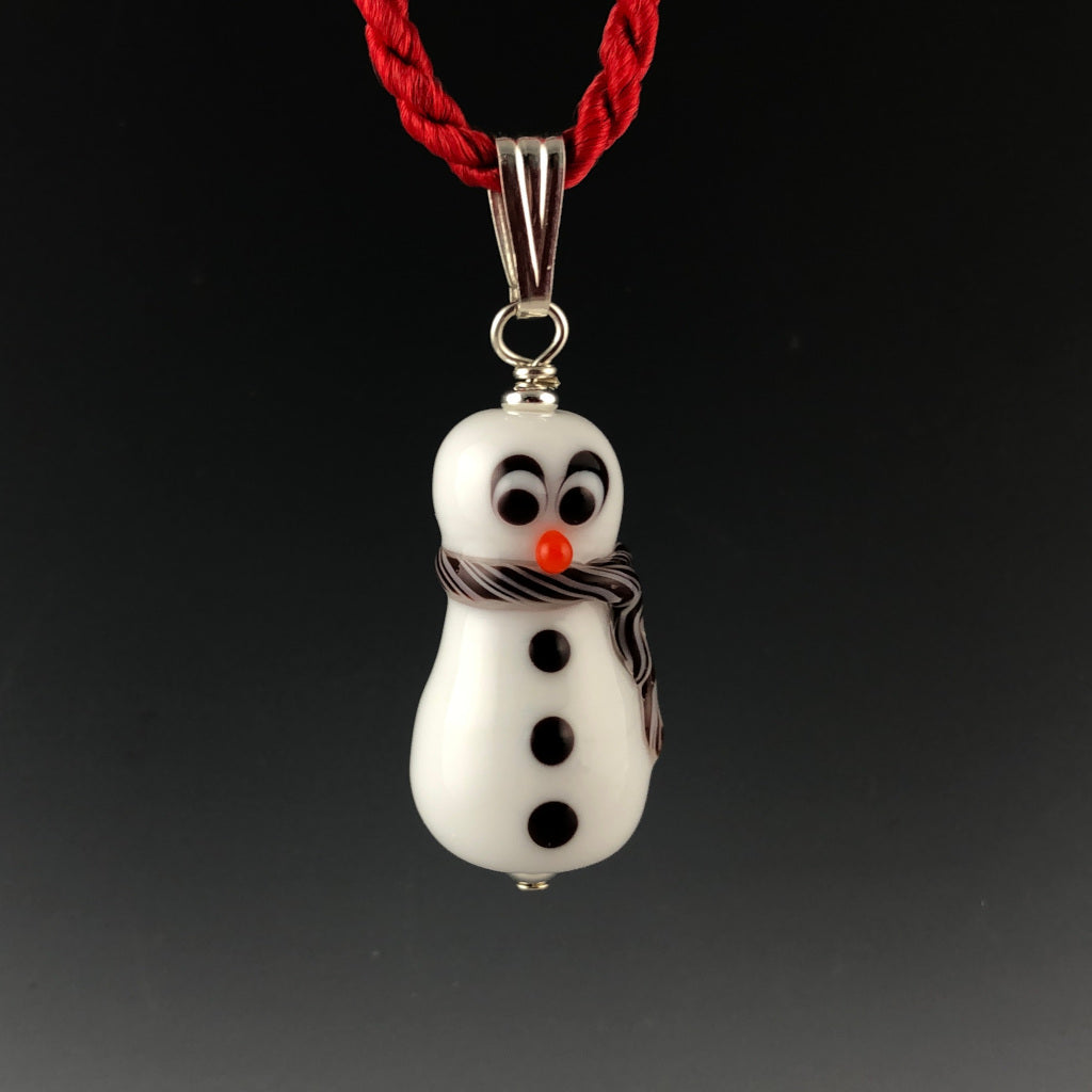 Glass snowman with surprized look with carrot colored nose, 3 black buttons, and white and red twisted scarf around neck and trails down his side. The snowman is on a sterling silver bail on a twisted red satin cord necklace.