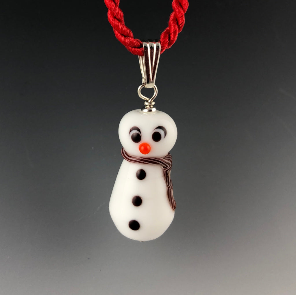 Holiday glass snowman pendant with a cross-eyed looking at his carrot colored nose, 3 black buttons, and white and red twisted scarf around neck and trails down his side. The snowman is on a sterling silver triangular bail on a twisted red satin cord necklace. Ready for Christmas!