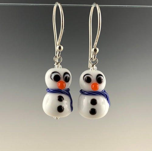 Petite snowmen earrings made of white glass with black and white eyes that look surprized. Each has 2 black buttons down their fronts and orange noses. They have blue and white glass scarves wrapped around their necks. They are on sterling silver ear wires. Zoom ready!