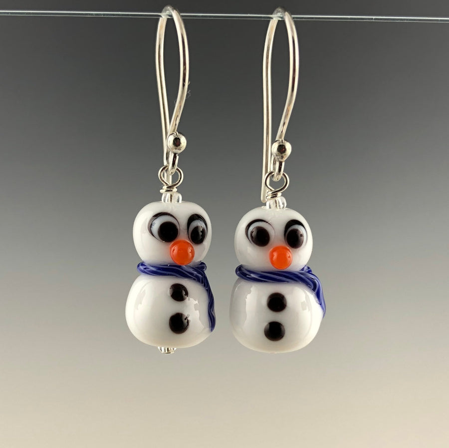 Petite snowmen earrings made of white glass with black and white eyes that look surprized. Each has 2 black buttons down their fronts and orange noses. They have blue and white glass scarves wrapped around their necks. They are on sterling silver ear wires. Zoom ready!
