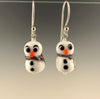 Petite snowmen earrings made of white glass with black and white eyes that look surprized. Each has 2 black buttons down their fronts and orange noses. They have blue and white glass scarves wrapped around their necks. They are on sterling silver ear wires. Ready for the winter holidays!