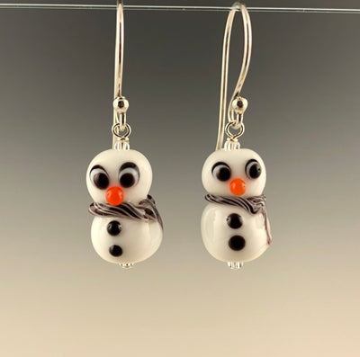 Petite snowmen earrings made of white glass with black and white eyes that look surprized. Each has 2 black buttons down their fronts and orange noses. They have blue and white glass scarves wrapped around their necks. They are on sterling silver ear wires. Ready for the winter holidays!